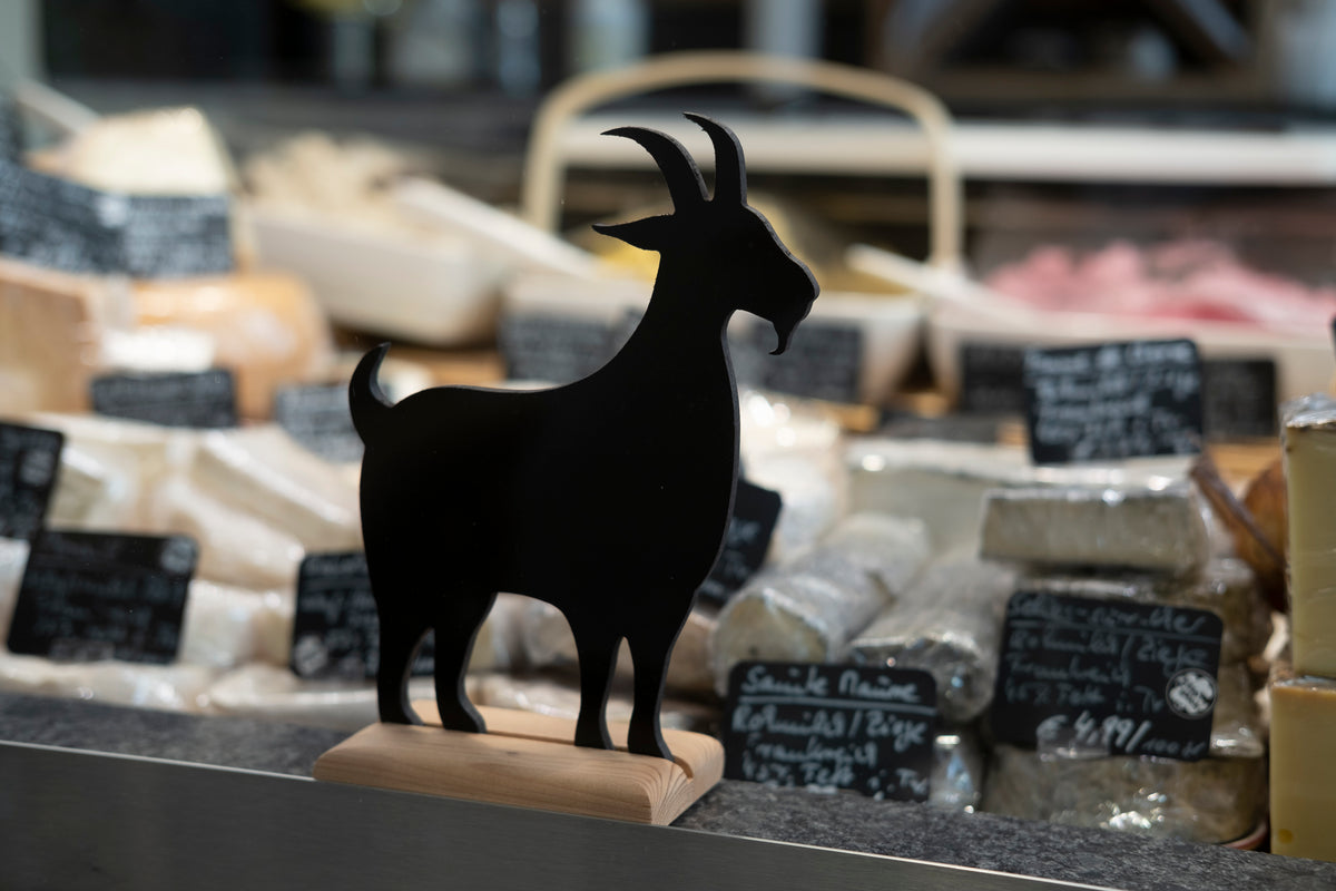Schwarze Ziegenfigur aus Holz auf der Käsetheke, dahinter Käsestücke mit Schildern